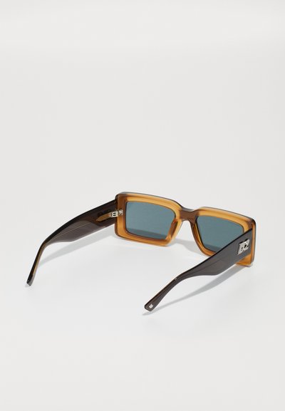 Amber frame sunglasses with dark brown arms, square lenses, and a metal accent logo on the temple. Lenses are tinted in a muted shade.