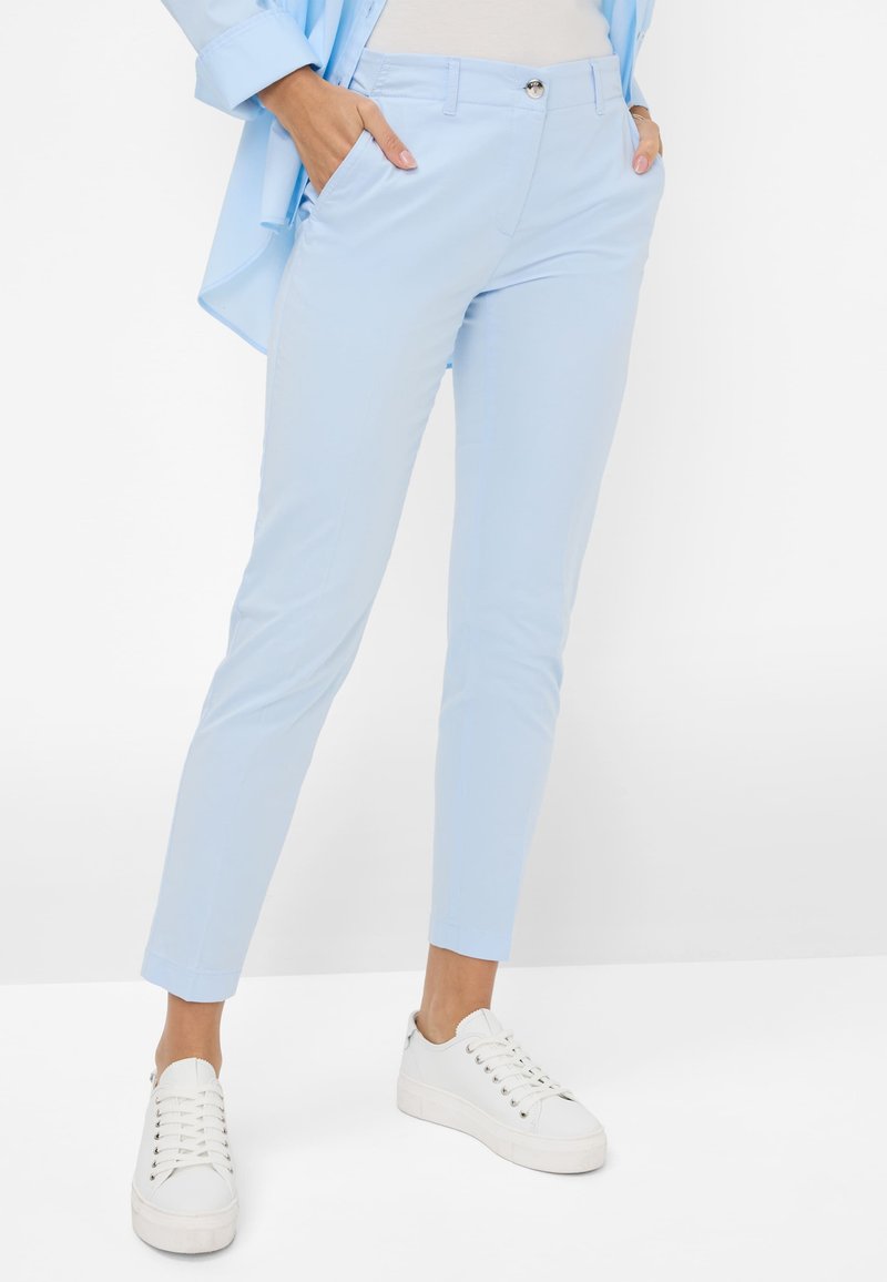 Lichtblauwe, slim-fit broek met rits- en knoopsluiting, zijzakken en een gladde textuur. Gecombineerd met witte sneakers.