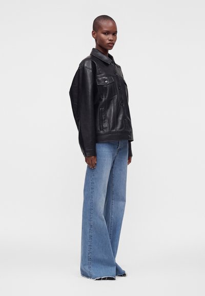 Personne de profil portant une veste en cuir noir et un jean bleu à jambes larges sur un fond uni clair.