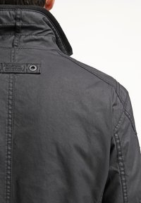 Vue arrière d'une personne portant une veste gris foncé avec des coutures visibles et une étiquette en tissu de marque près du col.