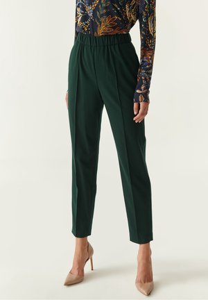 Femme portant un pantalon fuselé taille haute vert foncé et un haut à manches longues à motifs, associé à des talons aiguilles beiges à bout pointu.