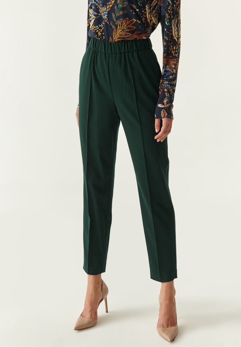Femme portant un pantalon fuselé taille haute vert foncé et un haut à manches longues à motifs, associé à des talons aiguilles beiges à bout pointu.