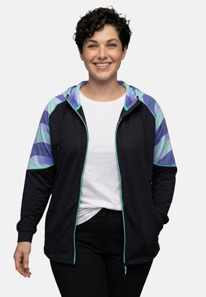 Uśmiechnięta osoba z krótkimi kręconymi włosami, ubrana w czarną bluzę z kapturem i zamkiem, z fioletowo-niebieskimi paskami na rękawach, na białej koszuli i czarnych spodniach.