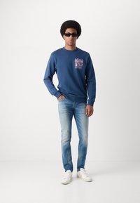Felpa blu con grafica rosa sul petto, abbinata a jeans azzurri e sneaker bianche. Il modello è in piedi con le mani nelle tasche.