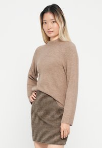 Brauner gestrickter Pullover mit hohem Ausschnitt und geripptem Saum, kombiniert mit einem kurzen Rock im Hahnentrittmuster in Dunkelbraun und Beige.