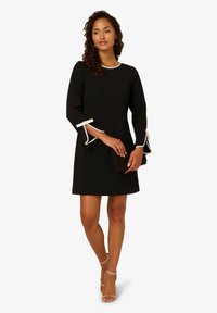 Černé šaty s tříčtvrtečními rukávy a bílým lemem na manžetách, doplněné černým psaníčkem a béžovými sandály na podpatku. Hladká textilie.