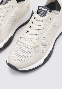 Zapatillas deportivas beige con parte superior de malla, detalles texturizados y suela blanca; cordones y logotipo en tonos contrastantes; diseño minimalista.