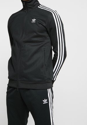 Personne portant un survêtement noir Adidas avec des bandes blanches sur les manches et le pantalon, debout devant un fond clair uni.