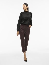 Schwarze Satinbluse mit hohem Kragen und gerafften Details; bordeauxfarbene, taillierte Hose mit schmaler Passform; schwarze spitze Schuhe mit goldenen Akzenten.