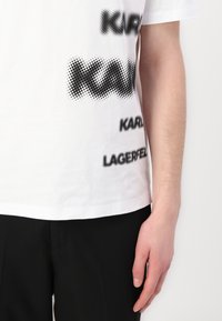 Bílá bavlněná tričko se černým textem různých písem a velikostí, včetně "KARL" a "LAGERFELD," v tučném písmu, s tečkovanými vzory.