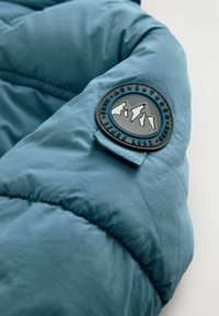Blau isolierte Jacke mit glänzender Oberflächenstruktur, ausgestattet mit einem runden Logo-Patch, das Berge zeigt. Sichtbare genähte Nähte.