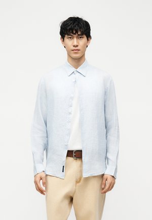 Jeune homme aux cheveux noirs bouclés portant une chemise bleu clair boutonnée sur un haut blanc, un pantalon beige et une ceinture marron, debout devant un fond blanc.