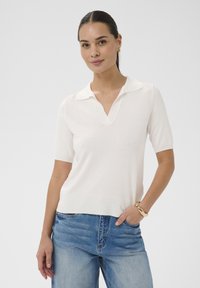 Femme portant un haut en maille blanc à manches courtes avec un col et un jean bleu, posant avec une main dans la poche sur un fond uni.