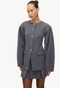 Veste grise sur mesure avec épaules structurées, boutons argentés et poches, assortie à une jupe plissée. Texture de tissu lisse.