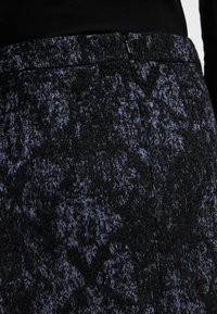 Gros plan sur une jupe en tissu texturé bleu foncé et noir avec une fermeture éclair latérale cachée, portée avec un haut noir.