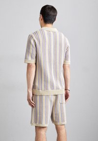 Gebreide polo en korte broek set in crème met blauwe en paarse strepen. Voorzien van een kraag, korte mouwen en geribbelde zomen. Textuurstof.