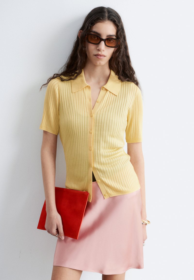 Femme aux longs cheveux bouclés portant des lunettes de soleil marron, un haut jaune à manches courtes boutonné, une jupe rose, tenant une pochette rouge.