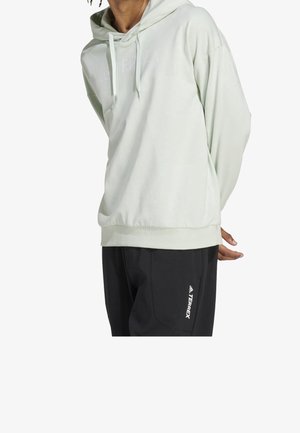 Sudadera verde claro con una textura suave, que cuenta con un bolsillo canguro, capucha con cordón y un logo sutil en la parte frontal. Combinada con pantalones negros.