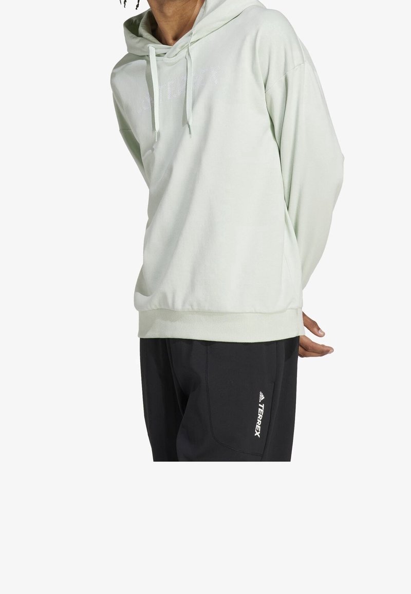 Hellgrüne Kapuzenjacke mit weichem Textil, ausgestattet mit einer Kängurutasche, einer Kapuze mit Kordelzug und einem dezenten Logo auf der Vorderseite. Kombiniert mit schwarzen Hosen.