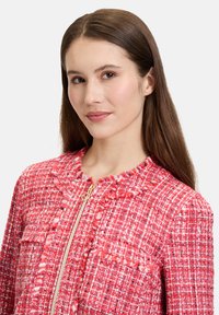 Czerwona kurtka o fakturowanej powierzchni z zapięciem na zamek, z okrągłym kołnierzem i dwiema kieszeniami na przodzie. Materiał ma tkany wzór z frędzlami.