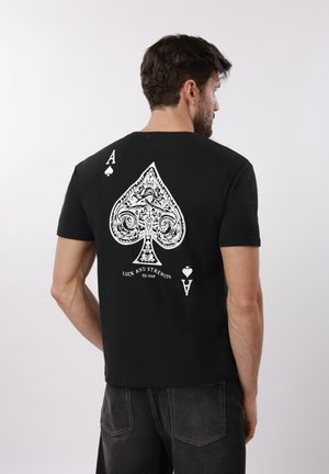 Hombre con camiseta negra con un diseño grande e intrincado de una carta de picas blanca y el texto "Suerte y Fuerza" en la parte trasera.