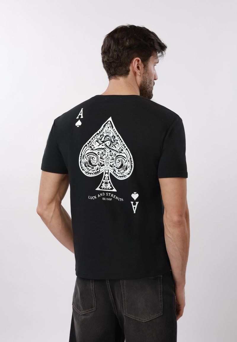 Moški, oblečen v črno majico s kratkimi rokavi, na hrbtu ima velik, zapleten bel znak srčka s tekstom "Sreča in moč".