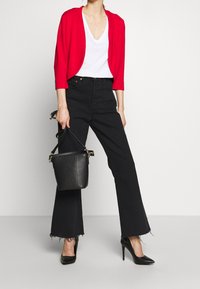 Cardigan rojo, camiseta blanca de escote en V, jeans negros acampanados con dobladillo deshilachado y tacones negros de punta. Sosteniendo un bolso negro tipo cubo con una correa.