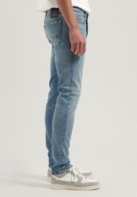 Jeans de mezclilla azul claro con corte ajustado, ligero desgaste, diseño de cinco bolsillos y parche con el logo en la cinturilla, combinados con zapatillas grises.