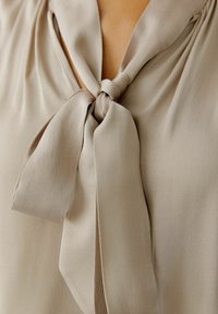 Beige Bluse mit einer gebundenen Schleife am Ausschnitt, aus glattem Material und fließendem Stoff. Der Stoff wirkt leicht und weich.