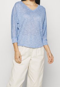 Hellblaues, langärmliges V-Ausschnitt-Oberteil aus strukturiertem Strickmaterial. Kombiniert mit beigen, locker sitzenden Hosen für einen lässigen Look.