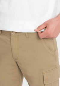 Pantaloni cargo beige con una texture liscia, dotati di chiusura con bottone, tasche laterali e accenti di cucitura. Ingrandimento della vestibilità vicino alla vita.