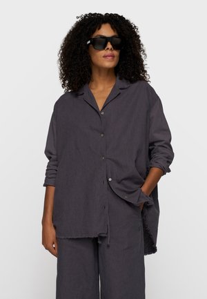 Vrouw met krullend haar draagt een oversized koolstofkleurig overhemd met knopen en bijpassende wijd uitlopende broek, aangevuld met grote zwarte zonnebrillen.