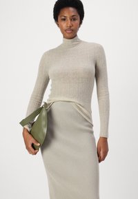 Beige geribbelde gebreide trui met een hoge hals, gecombineerd met een bijpassende geribbelde rok. Model houdt een groene leren handtas met een schouderband vast.