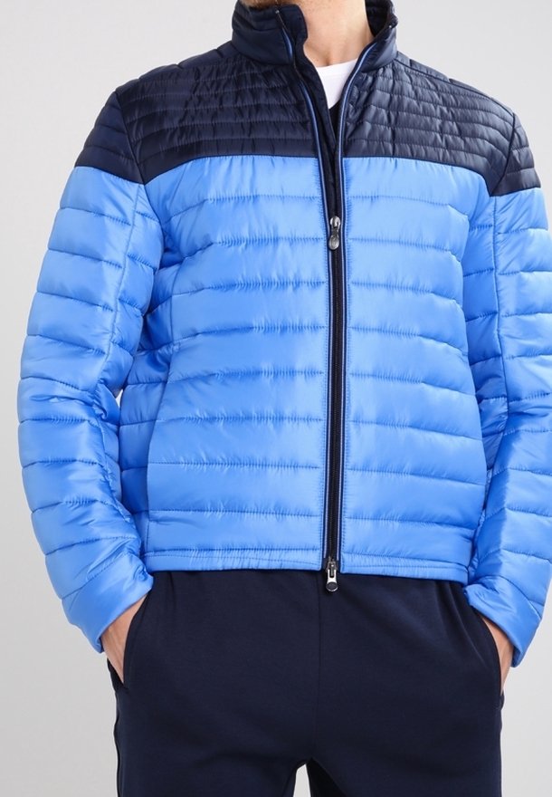 Blå puffjacka med en lätt design, horisontell sömnad, hög krage och kontrasterande mörkblå axlar; full dragkedjestängning.