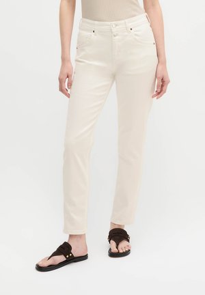 Frau trägt off-white Jeans mit geradem Bein und schwarzen flachen Sandalen, steht vor einem schlichten hellen Hintergrund.