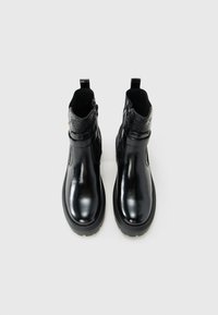 Une paire de bottines noires en cuir verni avec fermeture éclair latérale et détails gaufrés sur le dessus, présentées sur un fond blanc uni.