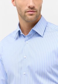 Camicia azzurro chiaro con sottili righe bianche, colletto classico, maniche lunghe e bottoni bianchi, caratterizzata da una texture del tessuto liscia.