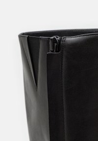 Botte en cuir noir à texture lisse, dotée d'une fermeture éclair latérale et d'un panneau élastique. Le design comprend un bord supérieur anguleux et net.