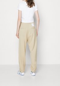 Beige wijde broek gemaakt van zachte stof, met twee achterzakken en een label op de tailleband. Gecombineerd met een effen witte top met korte mouwen.