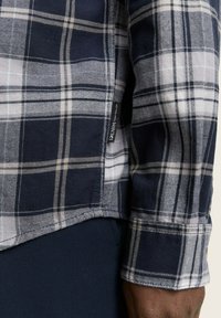 Koszula flanelowa w kratę w kolorze granatowym i białym, z kontrastowym dołem, z tkanym metkiem po lewej stronie w okolicy talii. Miękka w dotyku.