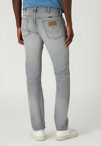 Vaqueros de mezclilla gris con un corte recto, que presentan bolsillos traseros con detalles de costura y un parche de cuero con la marca. Llevados con zapatillas blancas.