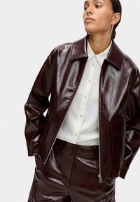 Bordeauxfarvet jakke i kunstlæder med blank finish, lynlåslukning foran og spids krave, kombineret med en hvid skjorte med knapper og matchende bukser.