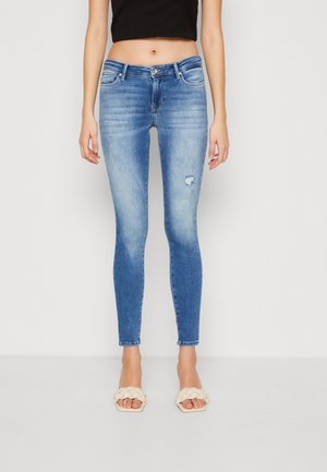 Jean bleu coupe slim taille mi-haute avec légère usure sur la cuisse droite, porté avec des sandales ouvertes à lanières tressées crème et un top court noir.