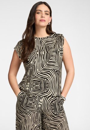 Kobieta nosząca dopasowany bezrękawnik i spodnie z czarno-beżowym abstrakcyjnym geometrycznym wzorem wijącym się, ręce w kieszeniach.