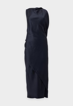 Femme portant une robe longue noire en satin sans manches avec un ourlet asymétrique et des sandales à talons beiges sur un fond clair uni.