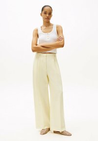 Top bianco senza maniche con scollatura rotonda e dettaglio con bottoni, abbinato a pantaloni ampi a vita alta di un leggero giallo e scarpe slip-on intrecciate.