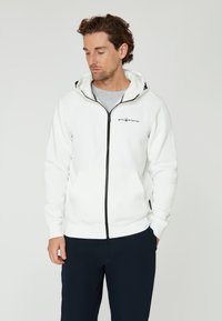 Sudadera blanca con cremallera y logo de la marca en el pecho. Cuenta con capucha y bolsillos laterales. Llevada sobre una camiseta gris.