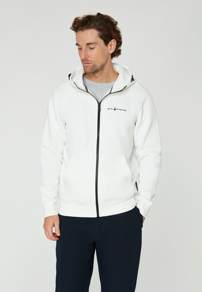 Sudadera blanca con cremallera y logo de la marca en el pecho. Cuenta con capucha y bolsillos laterales. Llevada sobre una camiseta gris.