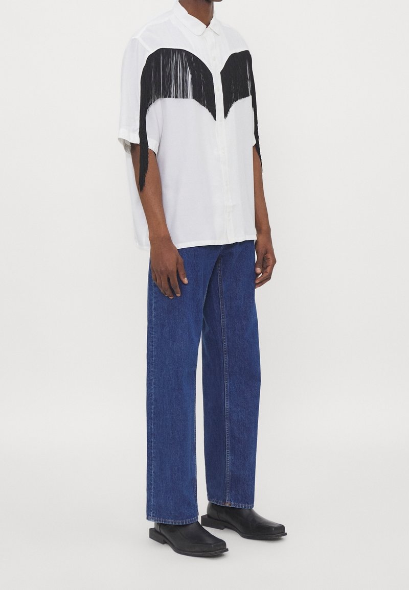 Camicia bianca a maniche corte con dettagli neri a frange sul petto, abbinata a pantaloni di jeans blu e stivaletti neri alla caviglia.