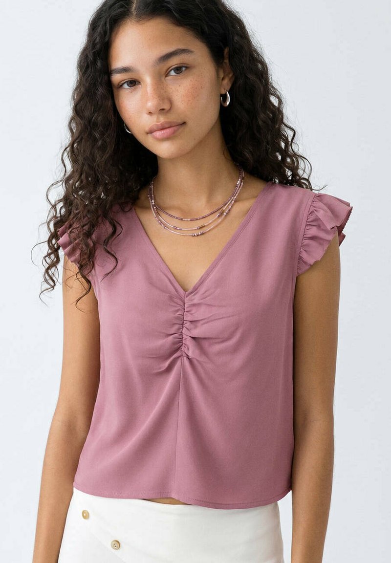 Jeune femme aux cheveux bouclés portant un débardeur mauve froncé et des colliers de perles superposés, debout devant un fond clair uni.